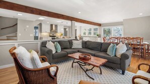 A spacious living room with a gray sectional sofa, a wooden coffee table, and wicker chairs. A kitchen with white cabinets and a large window is visible in the background. - Updated A-Frame w/ Game Room, Short Drive to Beach! (Barnstable)