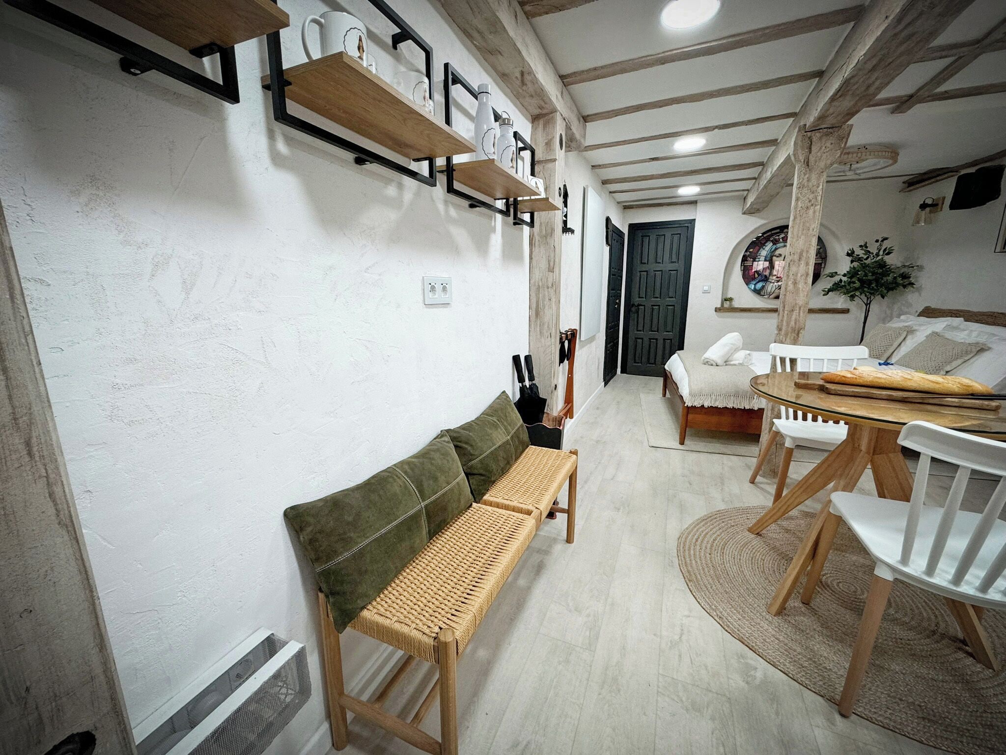 Une pièce chaleureuse avec un banc en bois, un lit recouvert de linge de maison blanc, une table à manger avec des chaises et une étagère murale agrémentée d’objets décoratifs.
