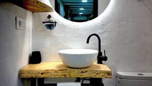 A bathroom with a wooden vanity, a white sink, a round mirror, and a small window. - St. John of the Cross Studio (San Sebastián de Garabandal)