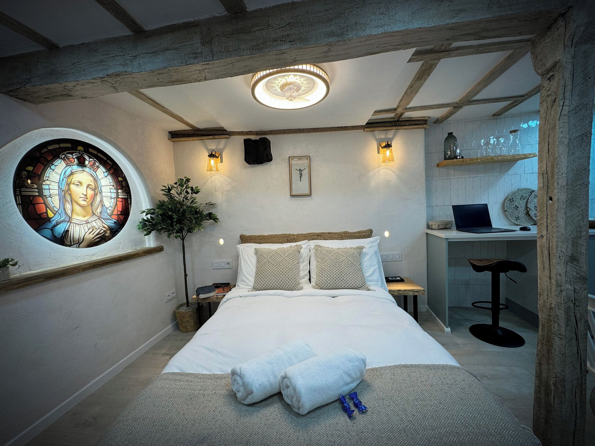 Une chambre à coucher avec un lit, un bureau et une chaise. Il y a une fenêtre ornée de vitraux et une plante.