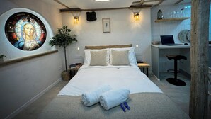 A bedroom with a bed, a desk, and a chair. There is a window with a stained glass design and a plant. - St. John of the Cross Studio (San Sebastián de Garabandal)