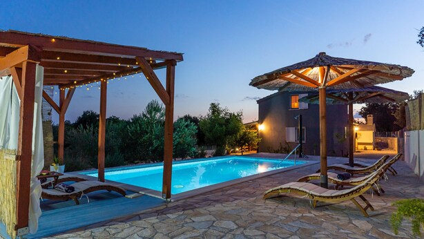 Area piscina con lettini e ombrelloni, circondata da pavimento in pietra e pergole in legno.
