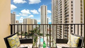 A balcony with wicker furniture, a table with a bottle, and a view of high-rise buildings and a swimming pool. - Ocean-view condo steps from Waikiki Beach (Honolulu)
