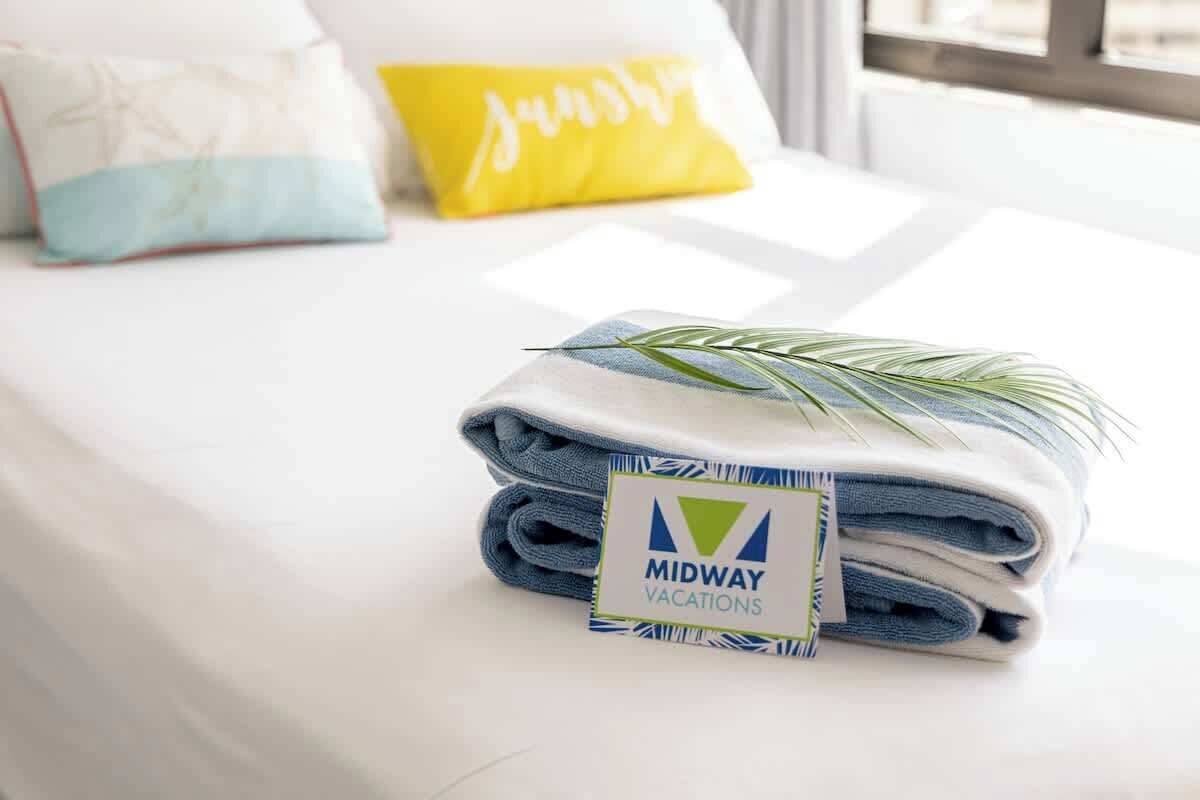 A neatly folded set of towels with a 'Midway Vacations' tag, a bed with white bedding, and a window in the background.