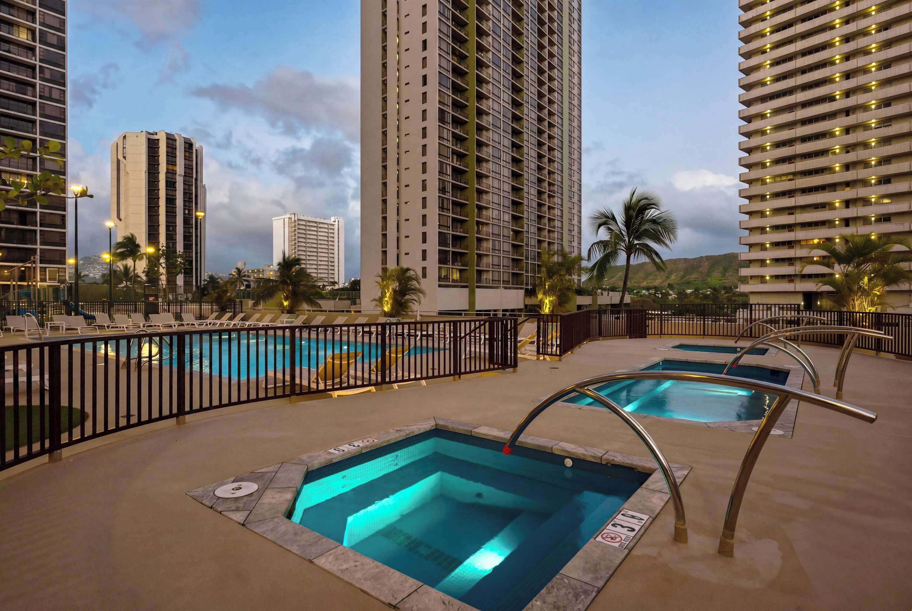 Un espace piscine d’hôtel comprenant plusieurs piscines, une clôture en métal noir et de grands immeubles en arrière-plan.