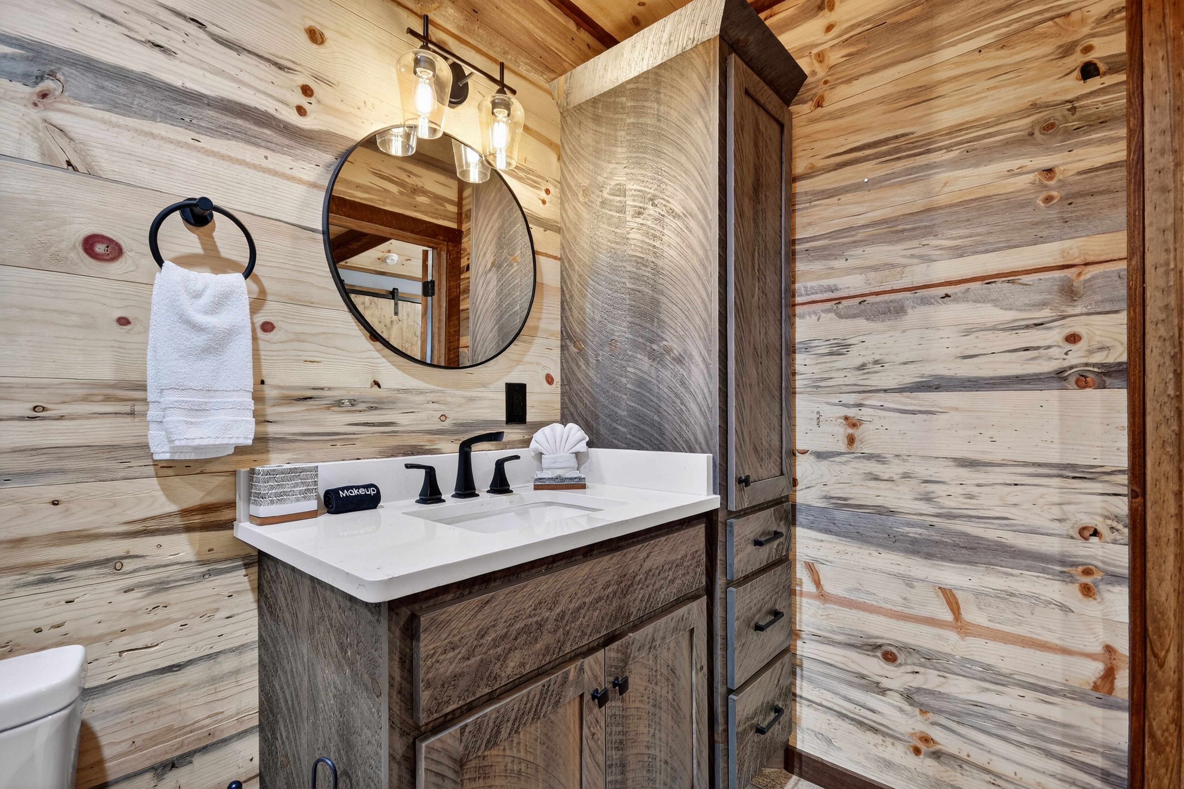 Un baño de cabaña de madera con un lavamanos blanco, grifo negro y un espejo redondo.