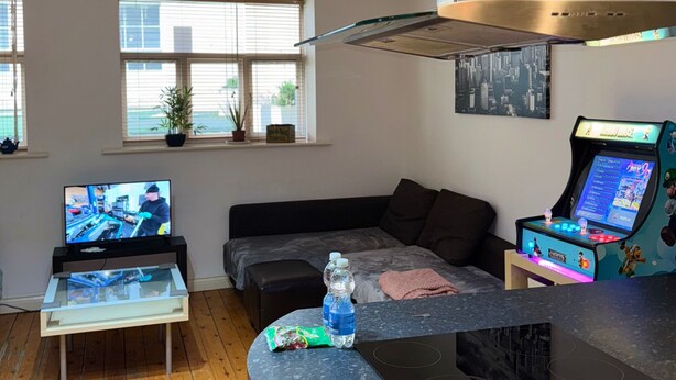 A modern living room with a kitchen area, including a stove, a gaming console, and a large window with blinds. - 1br-wifi-freesnacks-freeparking-petsok-retroarcade (Newcastle-upon-Tyne)