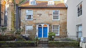 Un bâtiment traditionnel en pierre, avec une porte bleue, un petit jardin et une clôture en métal.