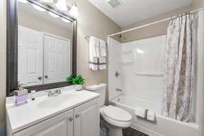 Une salle de bain avec un lavabo, des toilettes et une baignoire, tous de couleur blanche. Un miroir est placé au-dessus du lavabo, et un porte-serviettes contient deux serviettes.