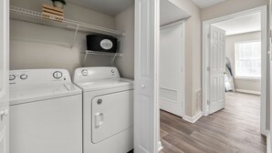 A laundry area with two white front-loading washers and dryers, a metal shelf with a potted plant, and a white door leading to another room. - Upper Westside Atlanta 2BR apartment with king beds, full kitchen, fast Wi-Fi, and free parking — close to dining, shopping, and MARTA. (Atlanta)
