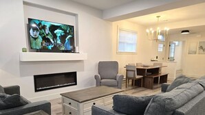 Una sala de estar moderna con un televisor de pantalla plana, sofás grises, una mesa de centro, una chimenea y un comedor con un candelabro.