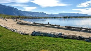 Vista sobre un lago con playa de arena, primer plano de césped y montañas al fondo.