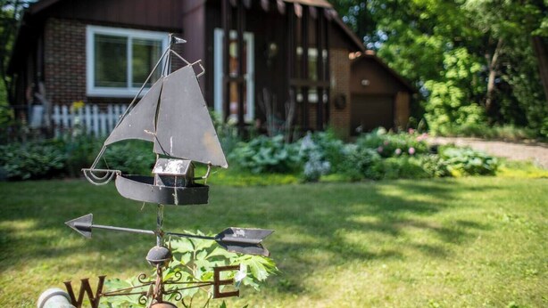 A weather vane with a sailboat design in a garden with a house in the background. - 1st flr nr Shaker, RTA rail, Cle Clinic & Univ. Ho (Akron)