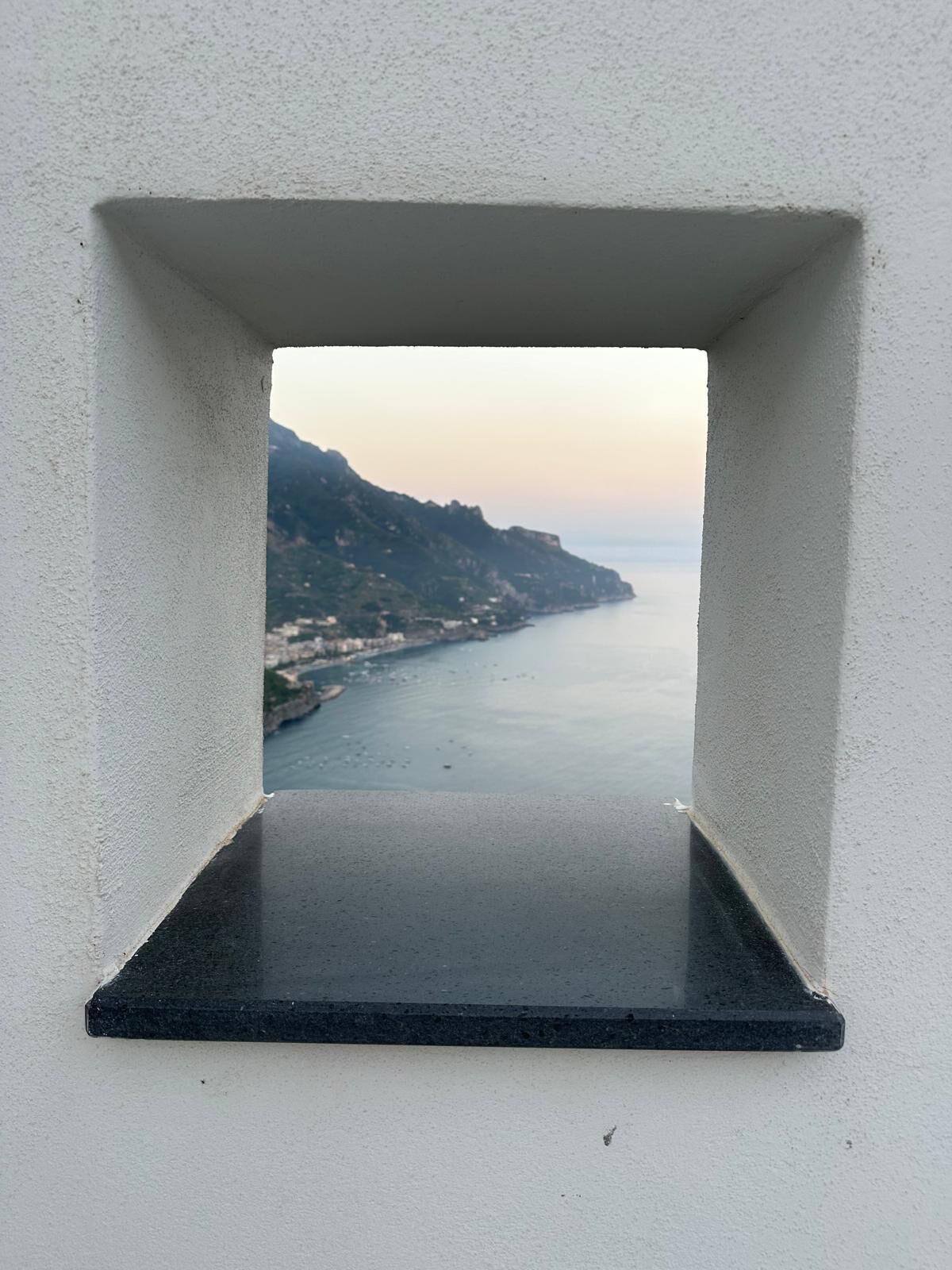 Vista attraverso una finestra con un davanzale nero, su una zona costiera con edifici e una catena montuosa.