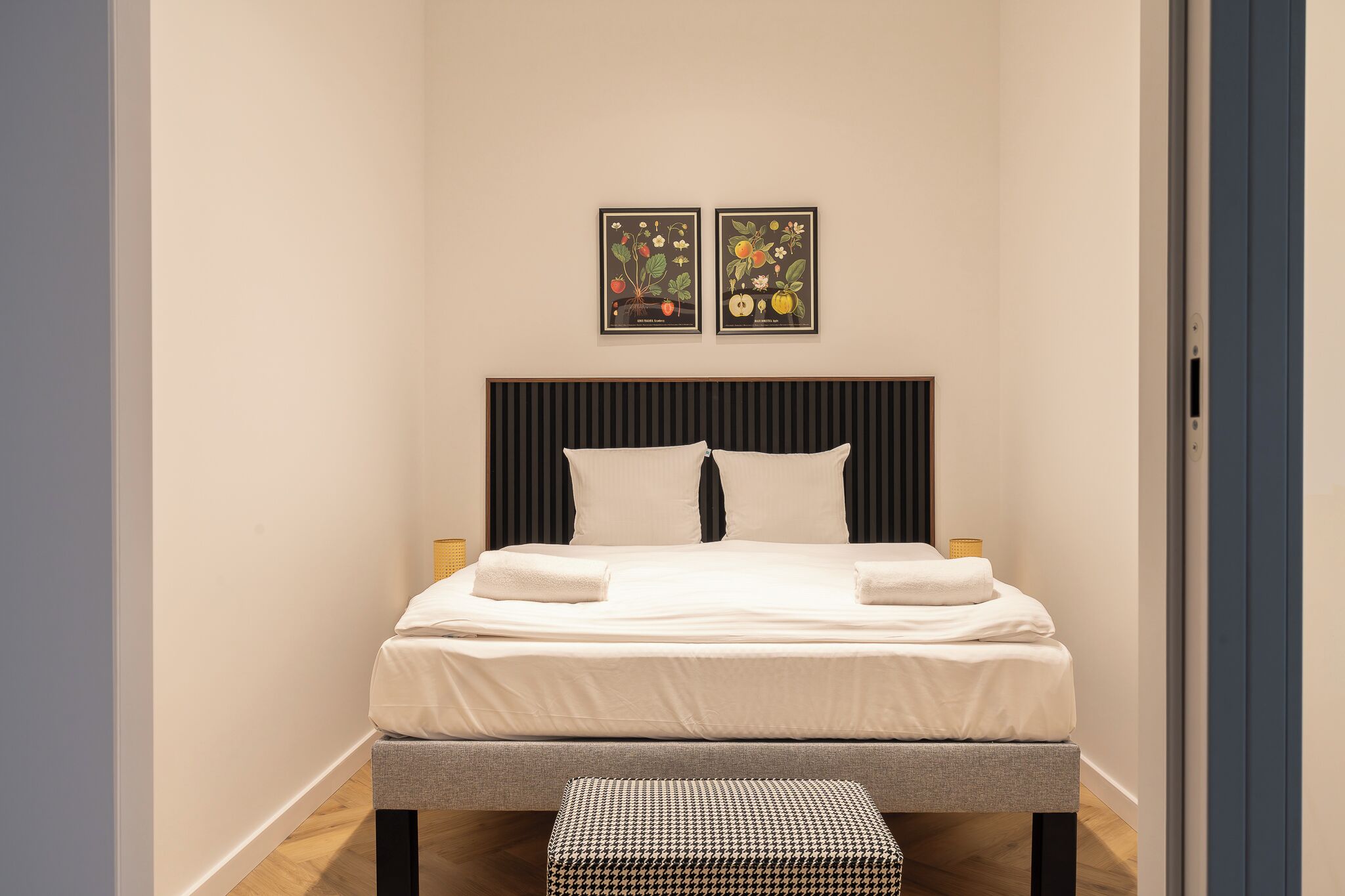 A neatly made bed with white linens and two pillows, a houndstooth bench, and two framed botanical prints on the wall.