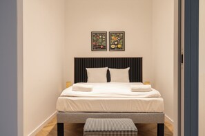 A neatly made bed with white linens and two pillows, a houndstooth bench, and two framed botanical prints on the wall. - RentPlanet - Przy Ogrodzie Botanicznym (Wroclaw)