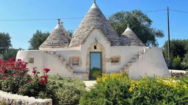Um pequeno edifício com cobertura de pedra, uma porta central e degraus que levam até ele, rodeado por vegetação e flores.