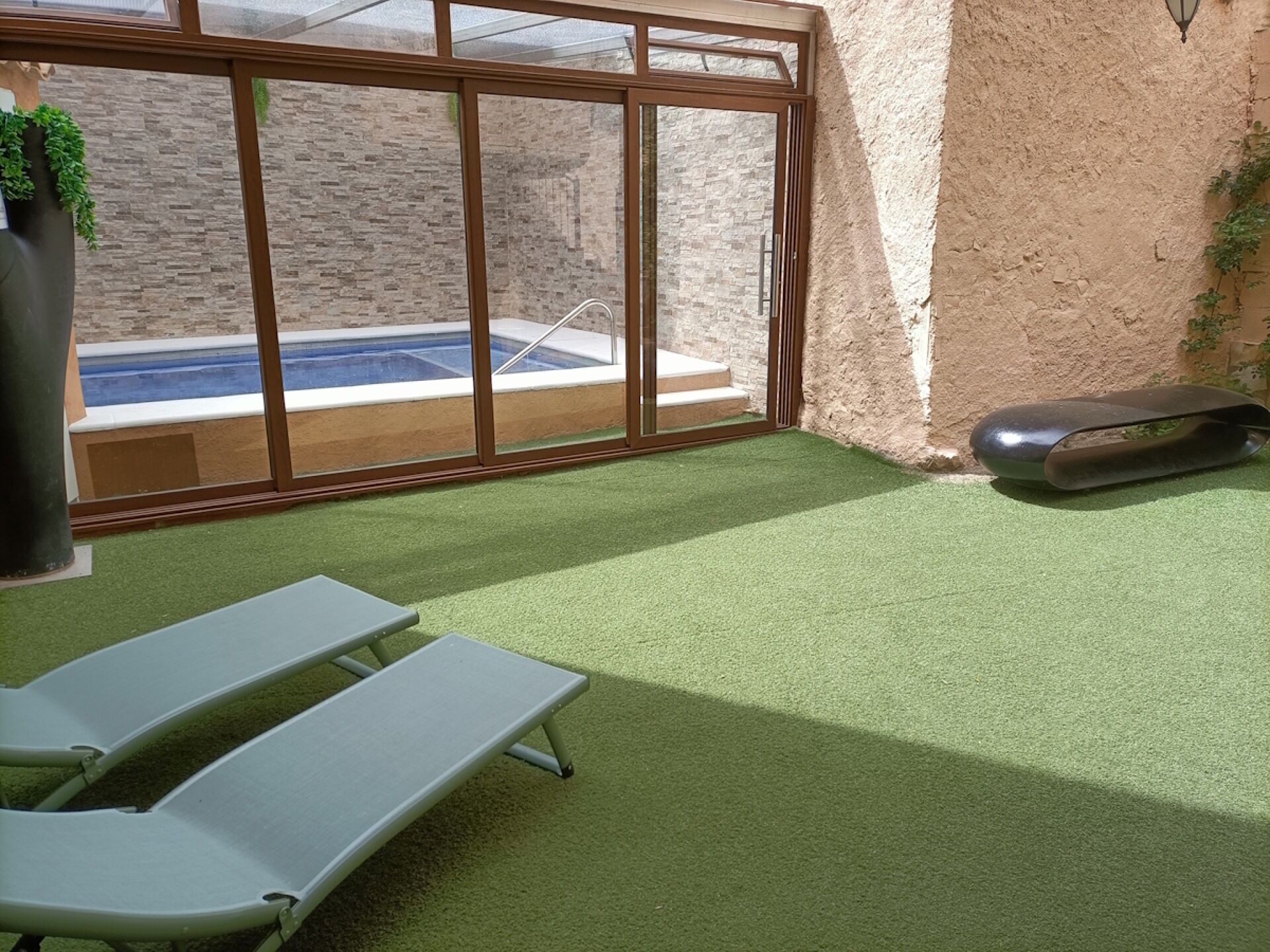 A sunlit patio with artificial grass, a pool, and a sliding glass door leading to a brick wall.
