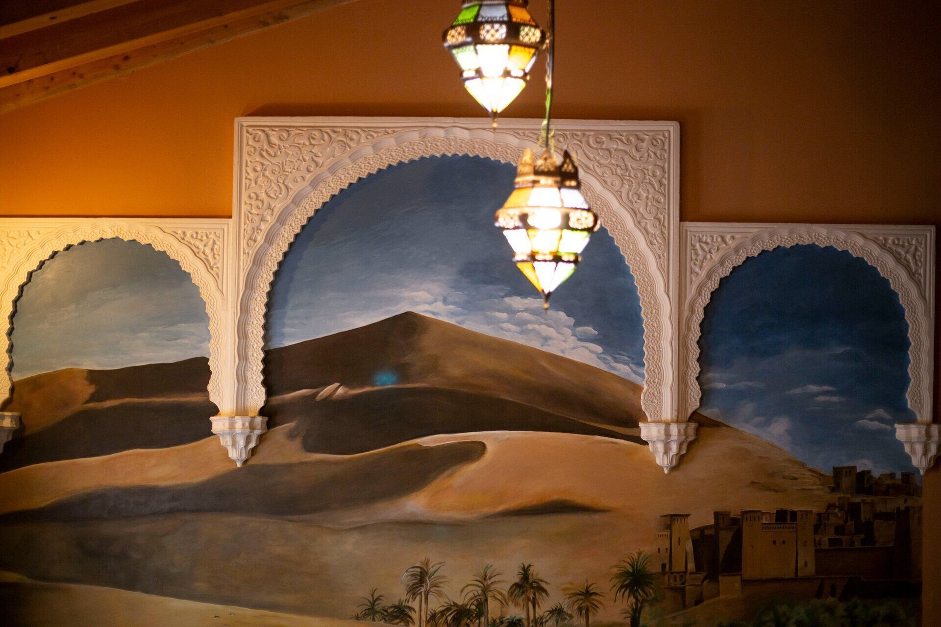 A mural of a desert landscape with palm trees and a cityscape, framed by ornate arches and a hanging lantern.