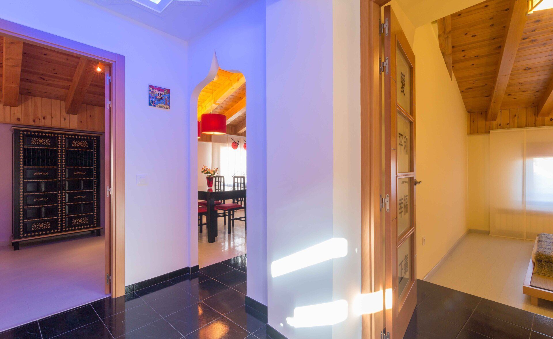 A hallway with a wooden ceiling, a black tiled floor, and a dining area visible through an open door.
