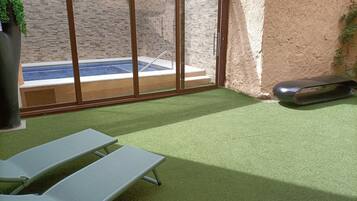 A sunlit patio with artificial grass, a pool, and a sliding glass door leading to a brick wall.