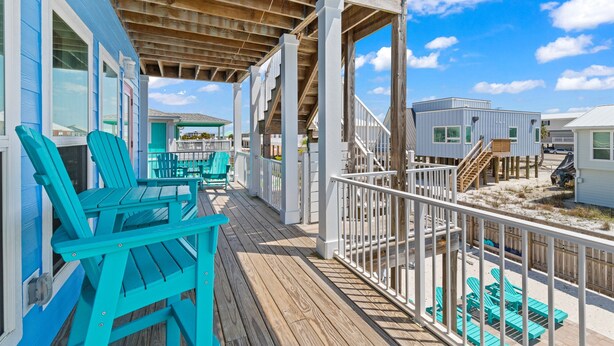 Eine Holzterrasse mit türkisfarbenen Stühlen und einem weißen Geländer mit Blick auf einen Strand.