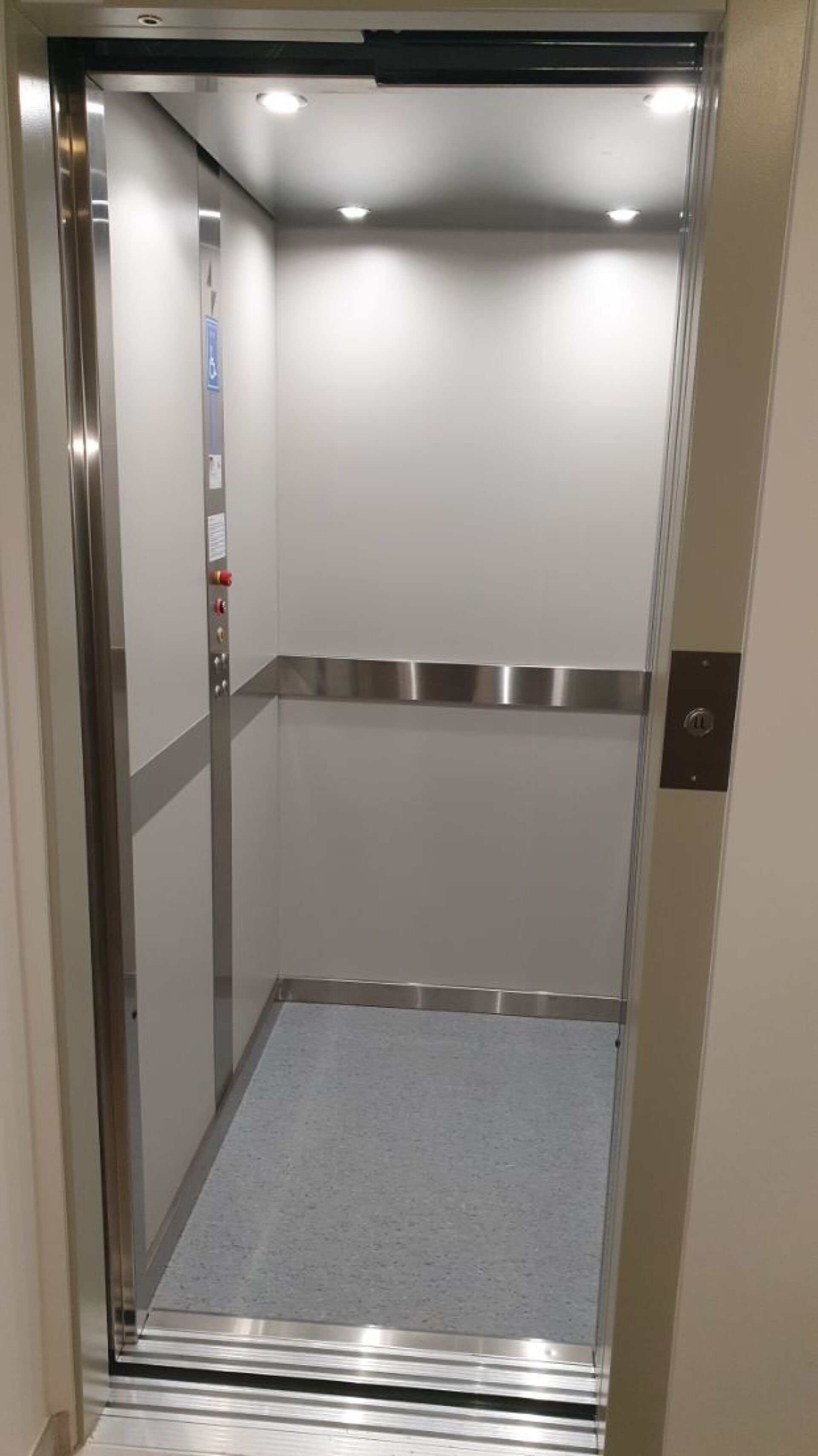 Un ascenseur aux parois métalliques, doté d’un tapis gris et d’un panneau de commande avec des boutons.