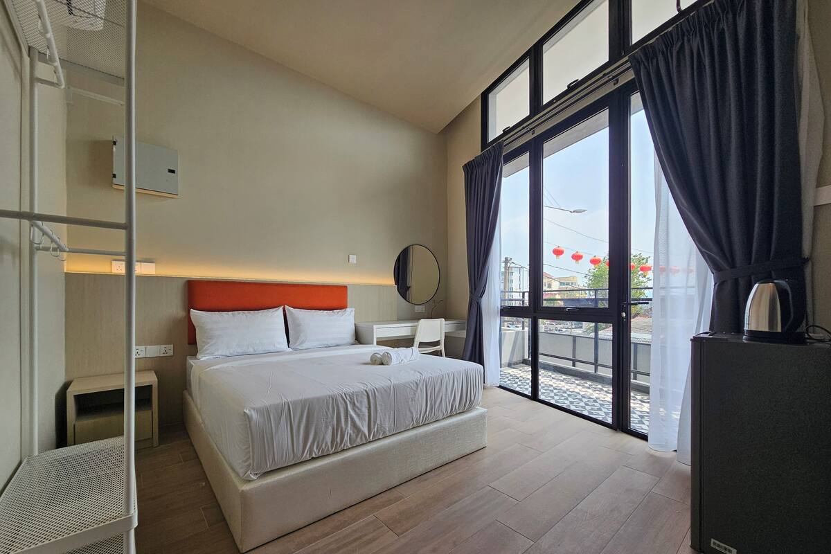 A hotel room with a large bed, a desk, a chair, a wardrobe, and a balcony with a view of the city.