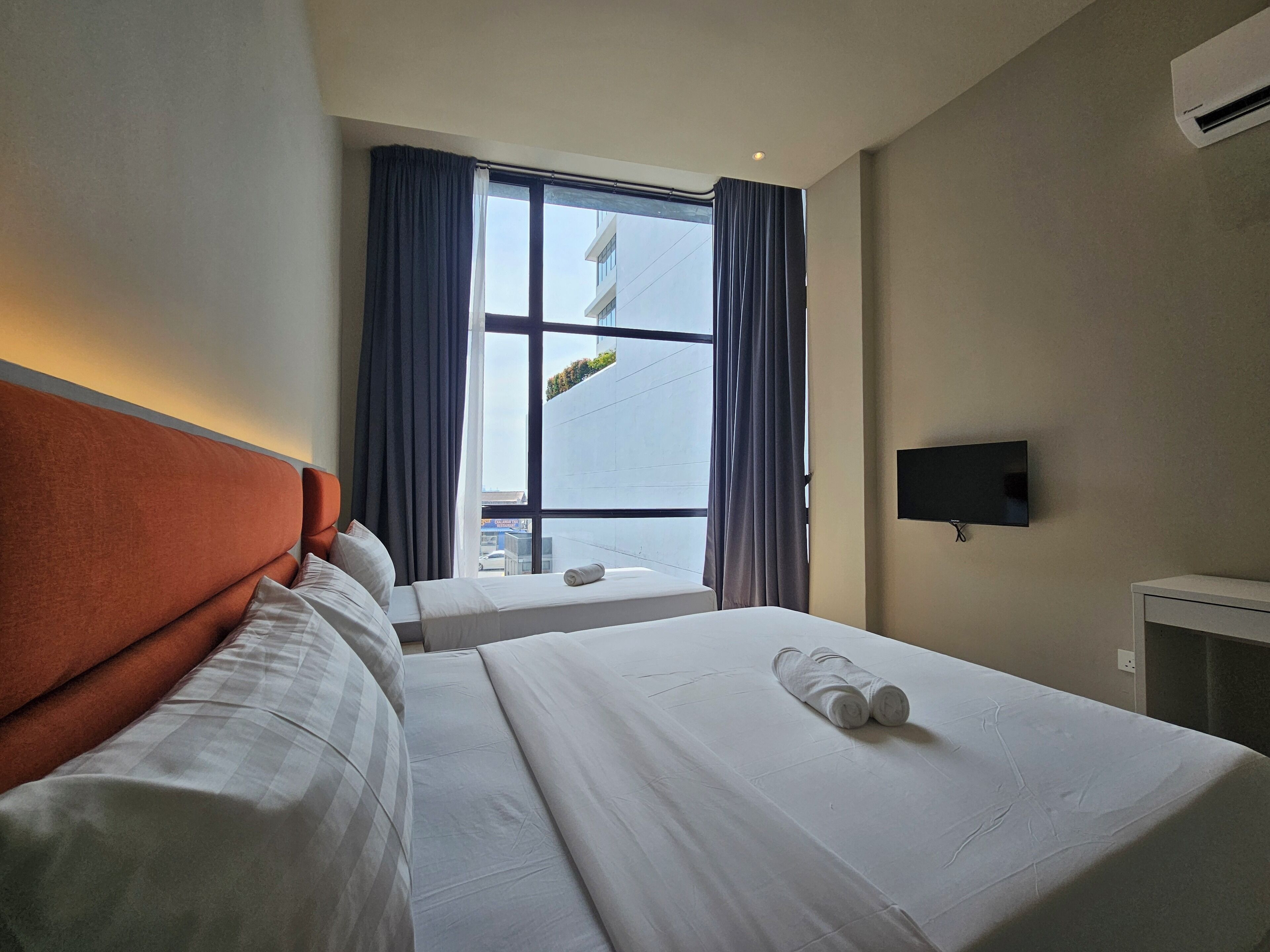 A hotel room with two beds, a large window with curtains, a flat-screen TV, and an air conditioning unit.
