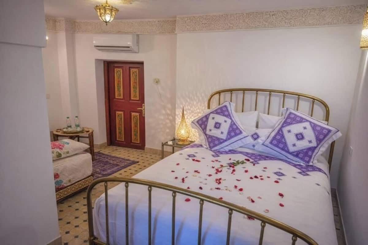 A bedroom with a bed, a nightstand, a lamp, a door, and a wall air conditioner.