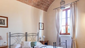 A bedroom with a brick ceiling, a metal bed frame, a wooden nightstand, a bedside table, a window with curtains, and a framed picture on the wall. - Ground floor apartment with one double bedroom (Toscana)