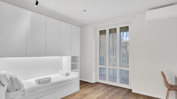 A modern, minimalist room with a white bed, built-in storage, and a large window with a view.