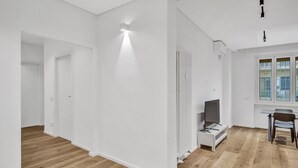 A spacious room with white walls, a wooden floor, a radiator, a television, and a window with blinds.