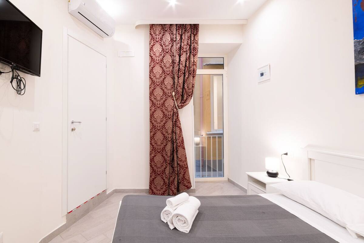 Una habitación de hotel moderna con una cama, una mesita de noche con lámpara, un ventanal con cortinas y un cuadro en la pared.