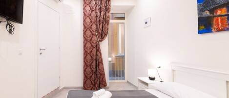 Ett modernt hotellrum med en säng, ett sängbord med en lampa, ett stort fönster med gardiner och en tavla på väggen.