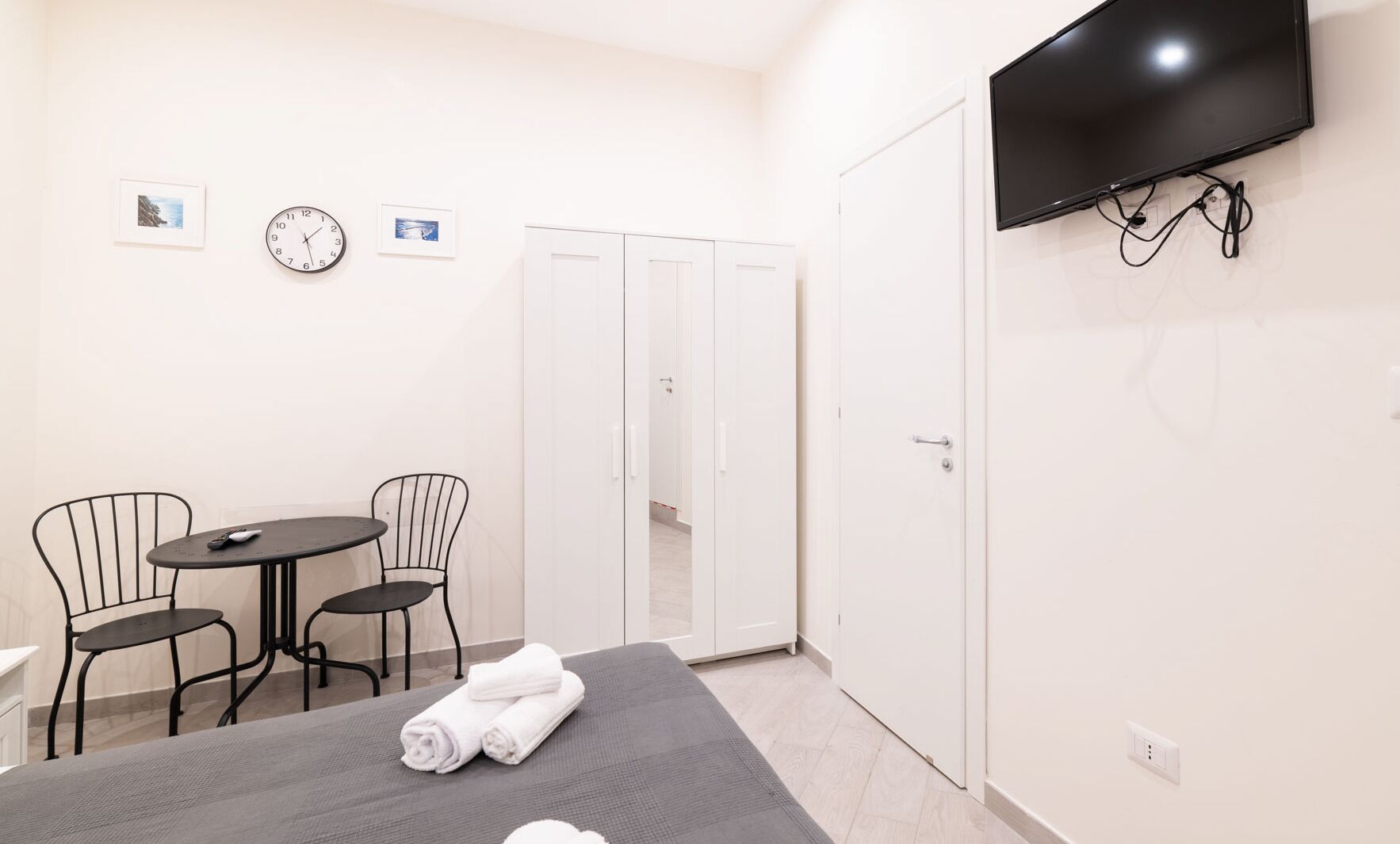 A hotel room with a bed, a TV mounted on the wall, a small table with two chairs, a clock, and a wardrobe.