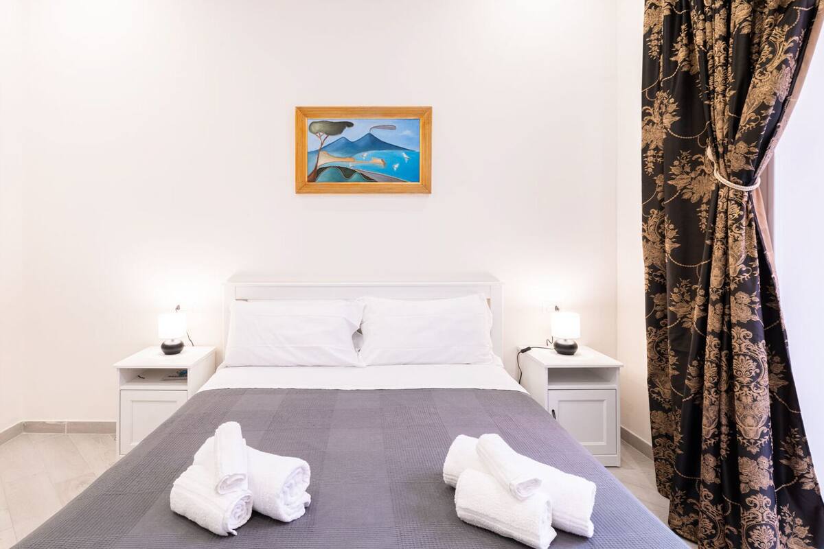 Una habitación de hotel con una cama, dos mesitas de noche con lámparas, un cuadro en la pared y una cortina.