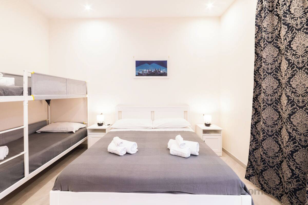 Una habitación con literas, una cama individual, dos mesitas de noche con lámparas y una cortina estampada.