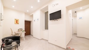 A hotel room with a flat-screen TV mounted on the wall, a desk with a chair, a small table with a lamp, and a door leading to another room. - Dimora Sergente Maggiore By DomusExtra (Naples)