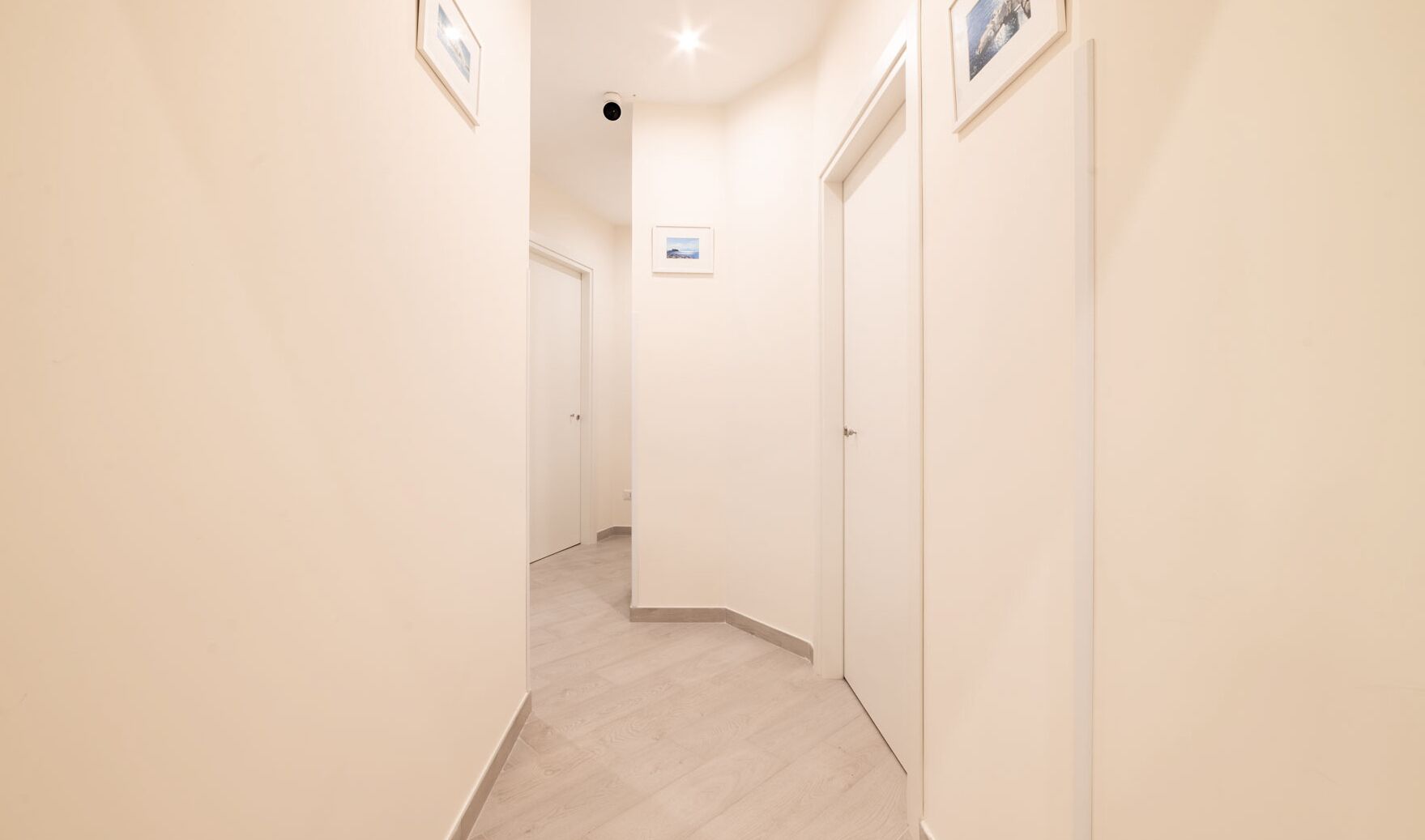 A narrow hallway with light-colored walls and wooden flooring, leading to a door at the end.