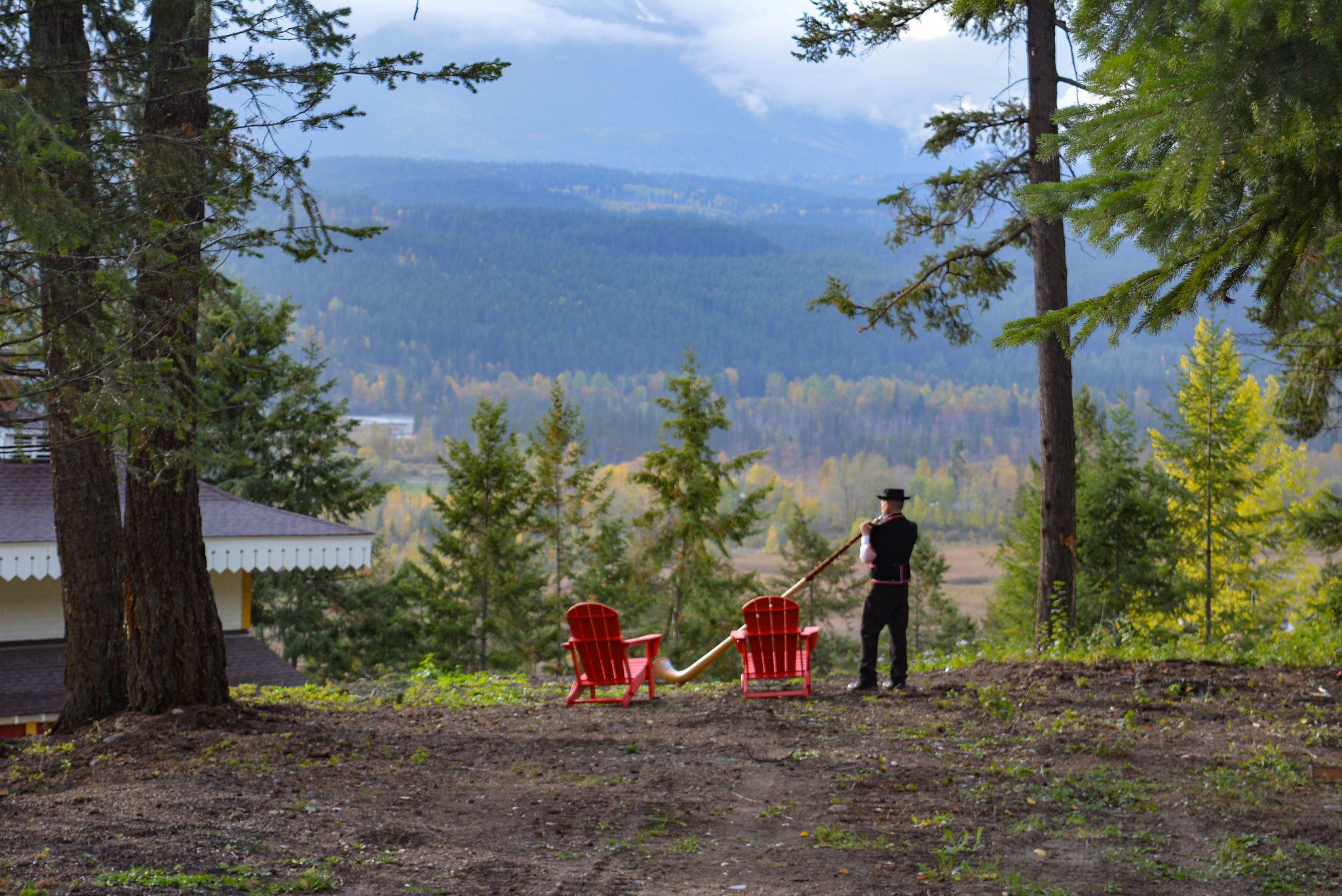 A person standing next to two red chairs and a shovel, overlooking a forested valley with a house in the background.