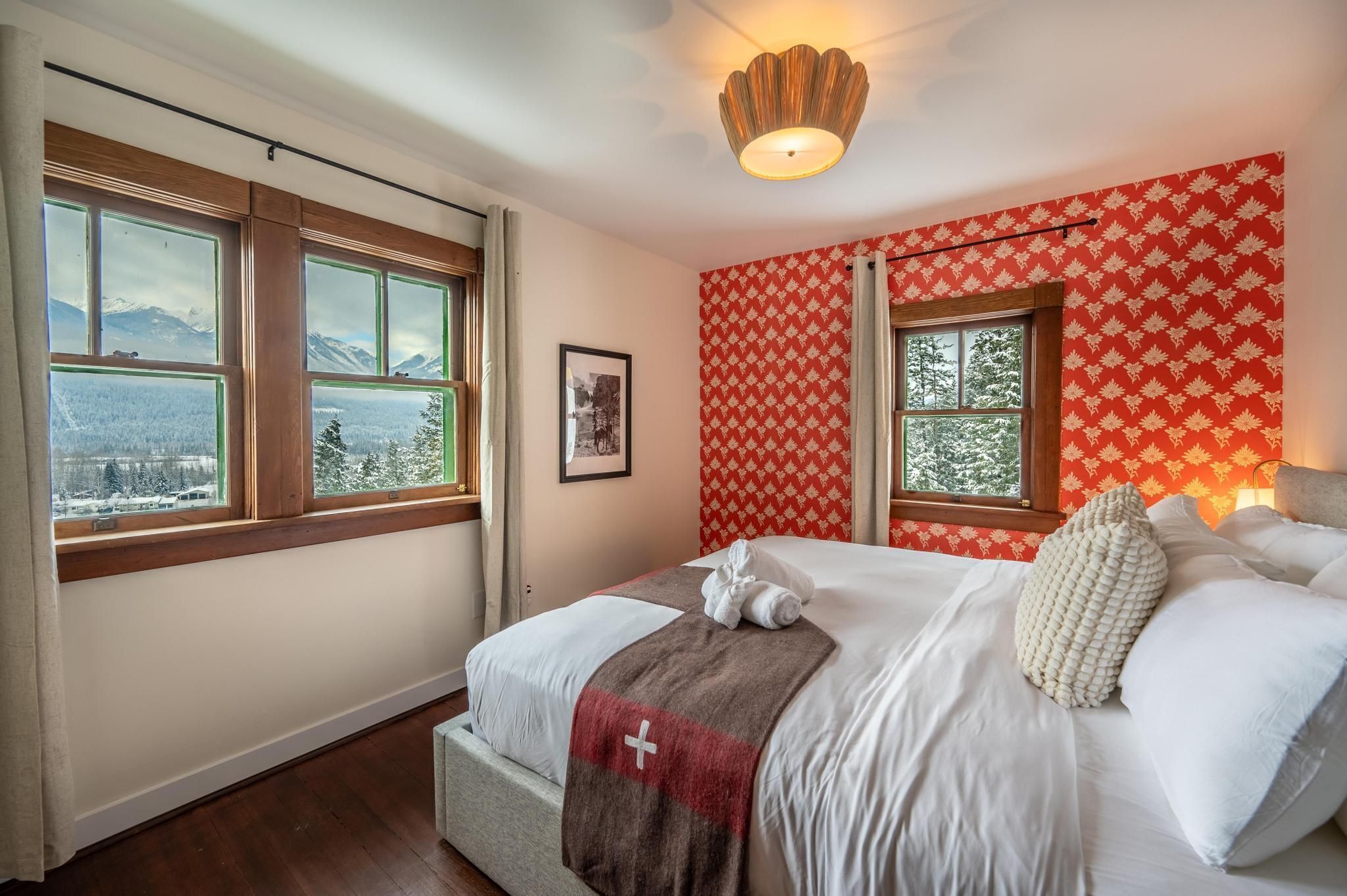Una habitación de hotel con una cama, dos ventanas, un cuadro enmarcado y una pared roja con estampado.