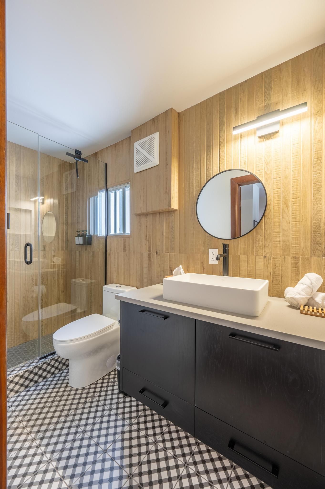 Een moderne badkamer met een glazen douchecabine, een wit toilet, een wastafelmeubel met een grote spiegel en een vloer met een ruitpatroon.