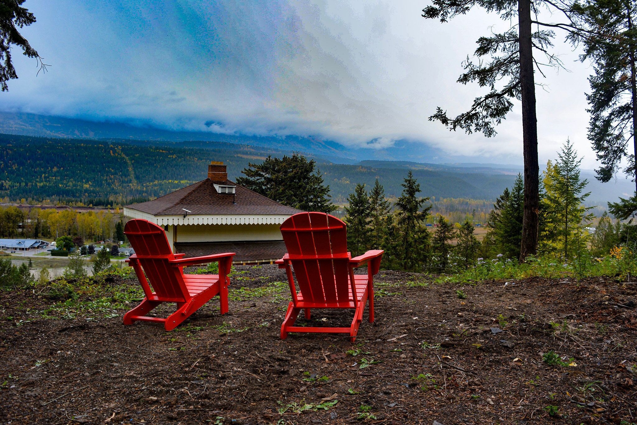 Two red Adirondack chairs facing a cabin with a view of a forested valley and mountains.