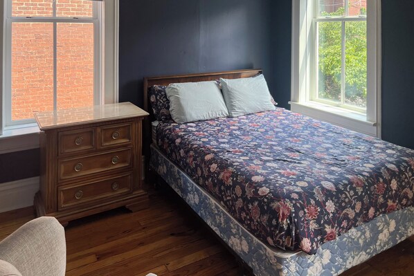 A bedroom with a wooden dresser, a floral bedspread, and a view of a brick wall through a window.