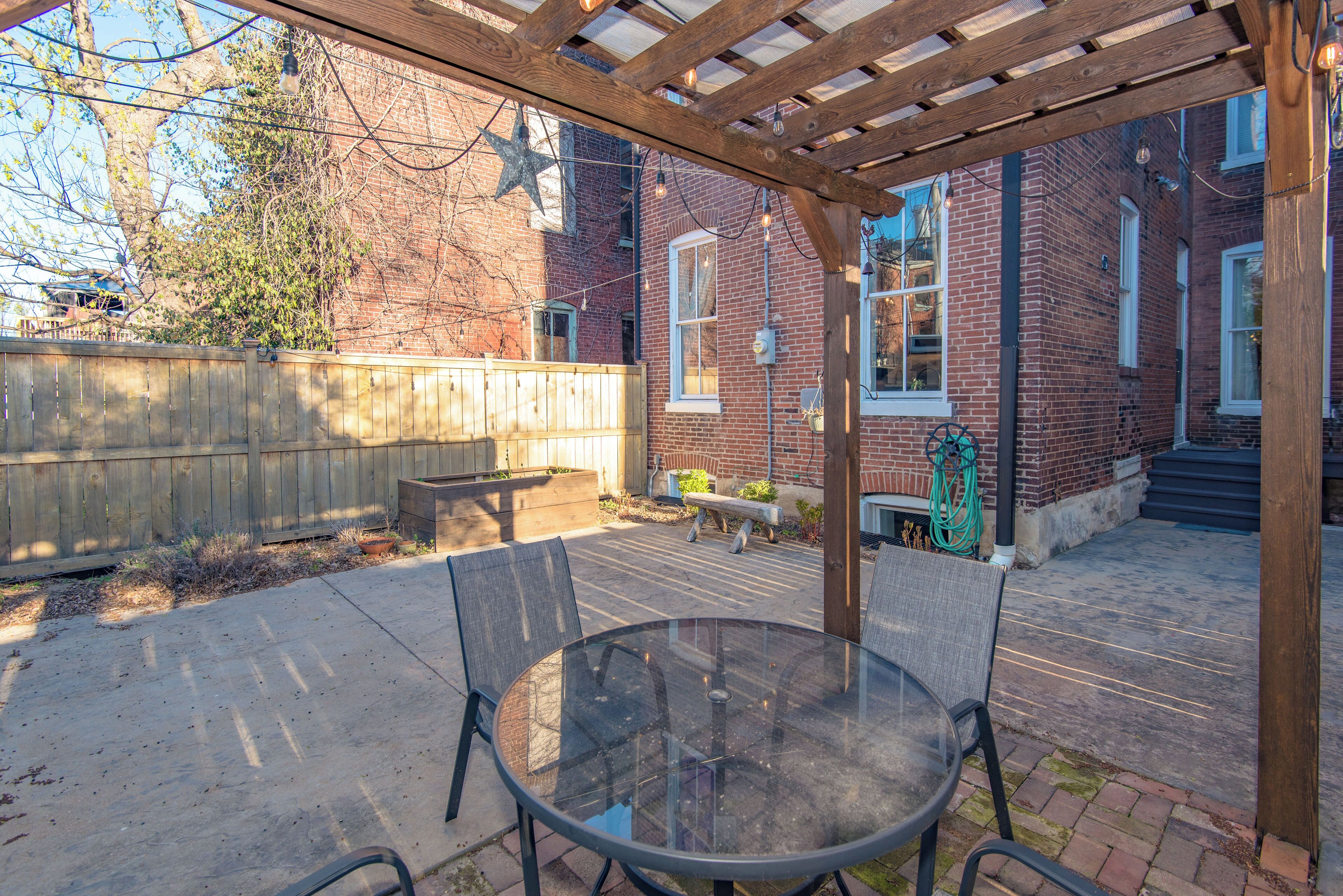 Une terrasse couverte avec une table et des chaises en verre, une pergola en bois et, au fond, un bâtiment en briques.