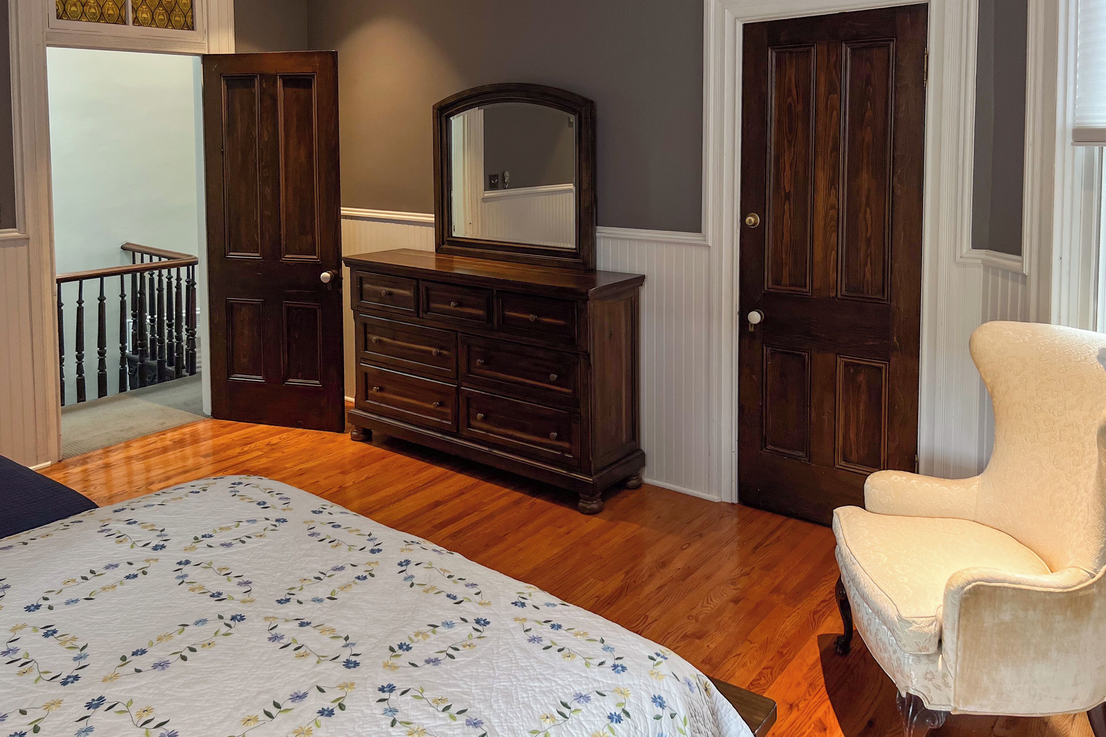 A bedroom with a wooden dresser, a chair, a bed with a floral bedspread, and a door leading to a staircase.
