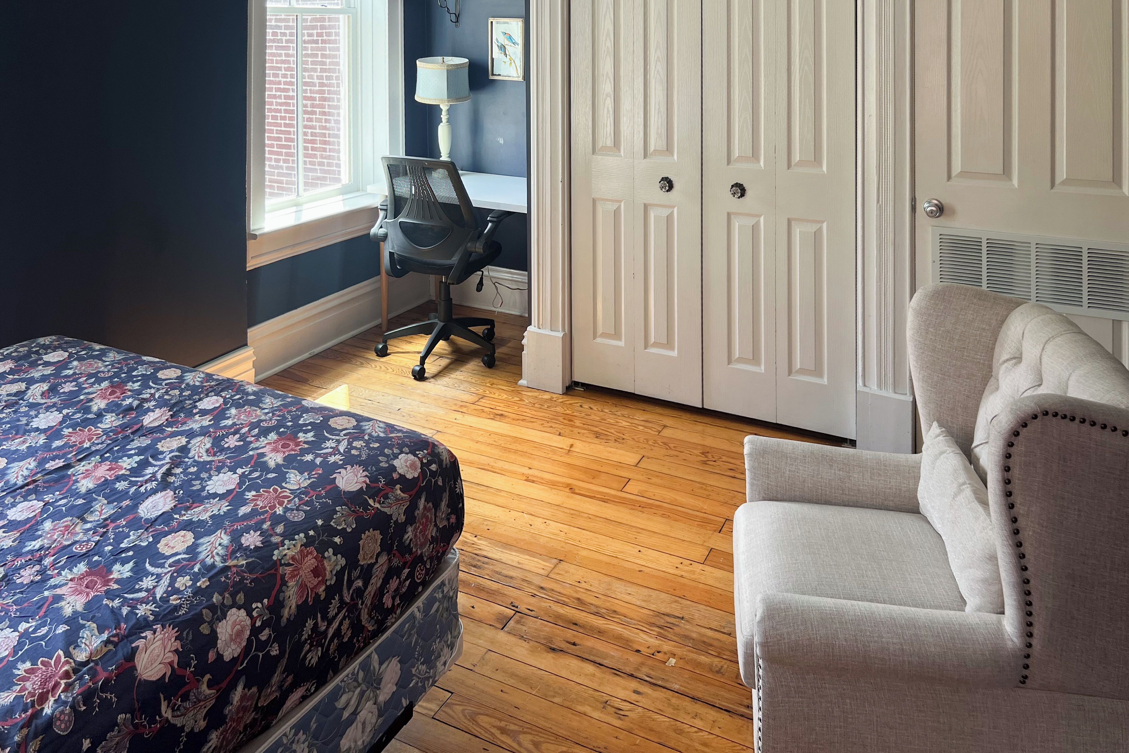 Une chambre à coucher avec une couvre-lit à motifs floraux, une chaise, un bureau avec une lampe et une armoire.
