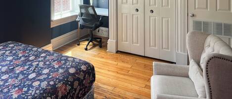 A bedroom with a floral bedspread, a chair, a desk with a lamp, and a wardrobe.