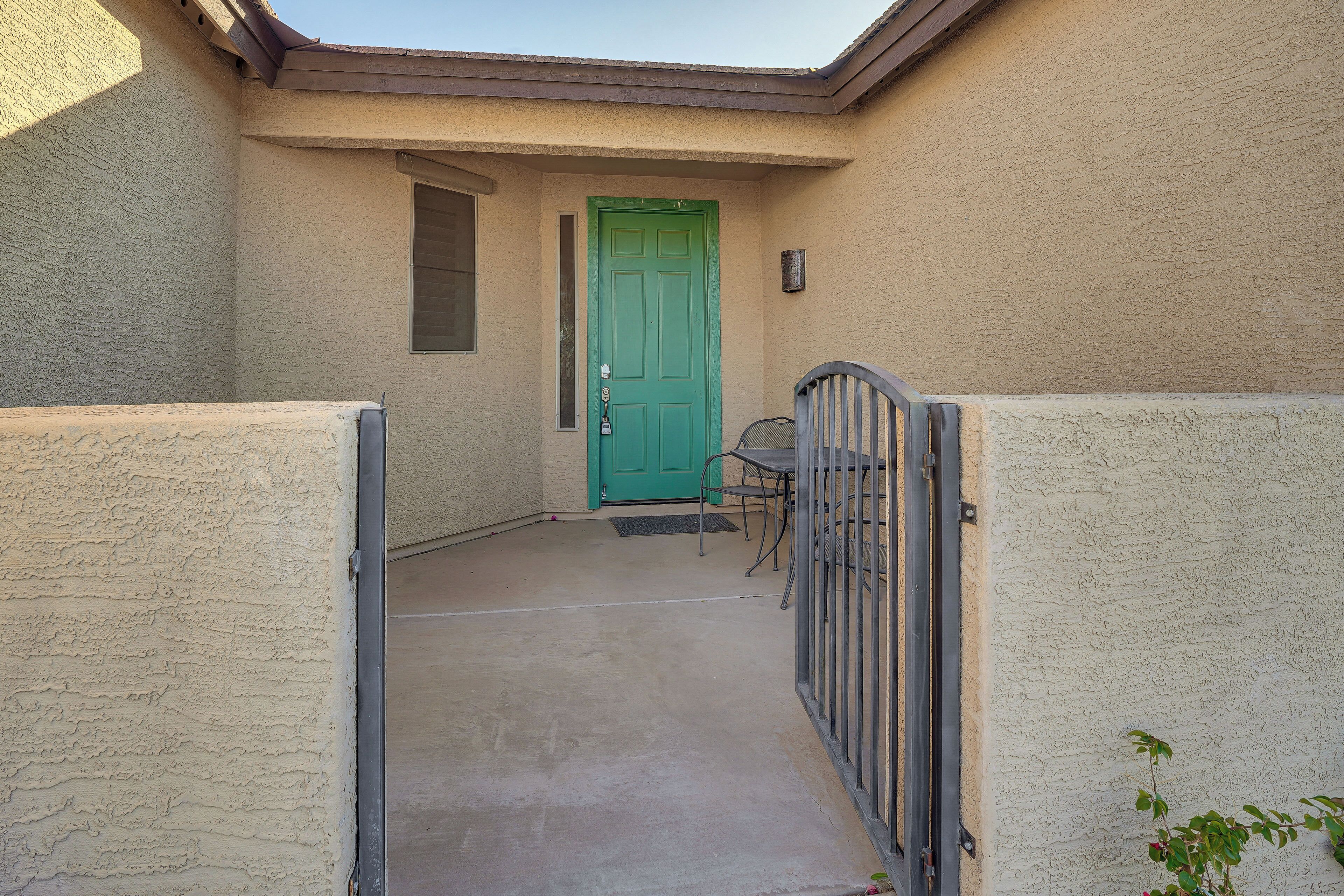 A narrow concrete pathway leading to a green door, with a metal gate on the right and a small table and chair in the center.